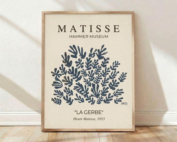 May include: Framed museum poster print leaning against white wall on light wood floor. Features Henri Matisse's 1953 "La Gerbe" artwork - dark blue botanical cutout shapes arranged in circular pattern on cream background. Simple wood frame, museum-style typography at top and bottom. Soft natural lighting creates diagonal shadows across white fabric backdrop.