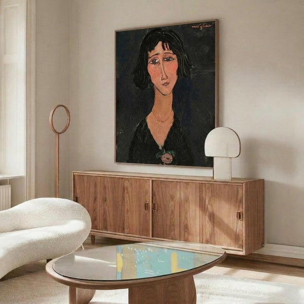 May include: Framed portrait print displayed above wooden sideboard in modern living room setting. Artwork shows woman with elongated face, dark hair, and black clothing in expressionist style with dark green background. White upholstered chair and glass coffee table visible in foreground, white table lamp on credenza.