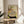 May include: Framed Japanese woodblock print showing a striped cat drinking from an ornate ceramic bowl, with pine branches and pinecones above. The vintage artwork features muted earth tones on cream background, displayed on a round wooden side table next to a beige upholstered chair in a bright, minimalist room with white walls and natural lighting.