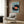 May include: Framed art print showing oversized vinyl record with orange center label surrounded by tiny people figures on light blue background, displayed above beige upholstered chair with geometric patterned pillow in modern living room with wood paneled walls, brass floor lamp, and dark round side table with open book