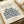May include: Beige framed art print featuring Henri Matisse's blue botanical cutout design from 1953, displayed on neutral wrapping paper with cream ribbon nearby. The minimalist poster shows organic leaf and branch patterns in navy blue against cream background, with museum-style text layout including "MATISSE HAMER MUSEUM" at top and artwork details below.