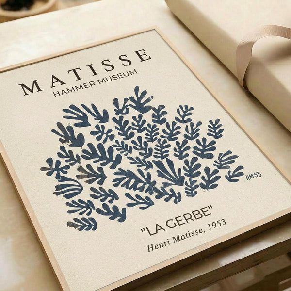 May include: Beige framed art print featuring Henri Matisse's blue botanical cutout design from 1953, displayed on neutral wrapping paper with cream ribbon nearby. The minimalist poster shows organic leaf and branch patterns in navy blue against cream background, with museum-style text layout including "MATISSE HAMER MUSEUM" at top and artwork details below.