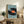 May include: Framed poster displaying a large black vinyl record with orange label surrounded by small people casting shadows on light blue background. Artwork shown in wooden frame on rustic sideboard next to fern plant, books, ceramic vase, and cream chair in modern living room setting.