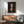 May include: Framed portrait artwork showing woman with dark hair and elongated features in expressionist style, displayed above wooden sideboard with table lamp, next to mid-century modern green upholstered chair with wooden arms, white walls with natural sunlight shadows