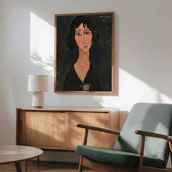 May include: Framed portrait artwork showing woman with dark hair and elongated features in expressionist style, displayed above wooden sideboard with table lamp, next to mid-century modern green upholstered chair with wooden arms, white walls with natural sunlight shadows