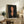 May include: Framed portrait print of a woman with black hair and elongated features in modernist style, displayed on rustic wooden console table alongside potted fern, ceramic vase, and books in neutral-toned living room setting with beige chair visible