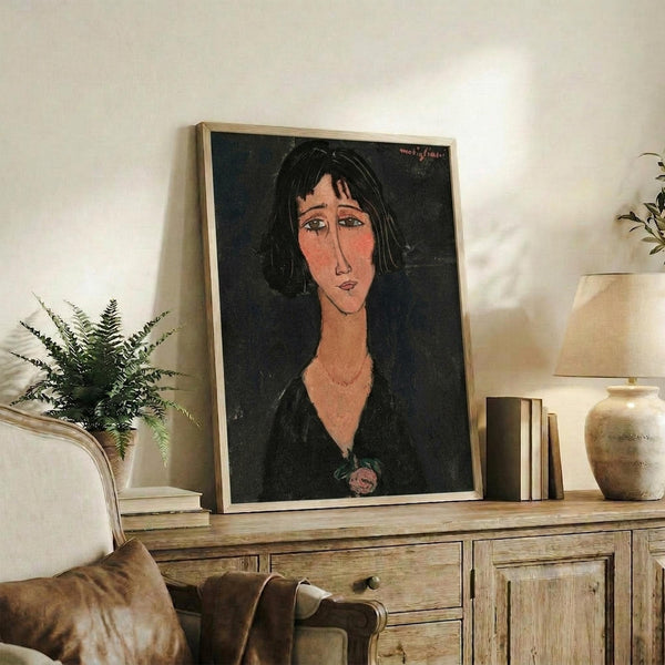 May include: Framed portrait print of a woman with black hair and elongated features in modernist style, displayed on rustic wooden console table alongside potted fern, ceramic vase, and books in neutral-toned living room setting with beige chair visible