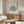May include: Framed poster with blue botanical leaf cutout design on cream background, displayed above wooden sideboard in modern living room. White sculptural chair and glass-top coffee table with abstract art visible. Warm natural lighting creates shadows across neutral-toned space with minimalist decor.
