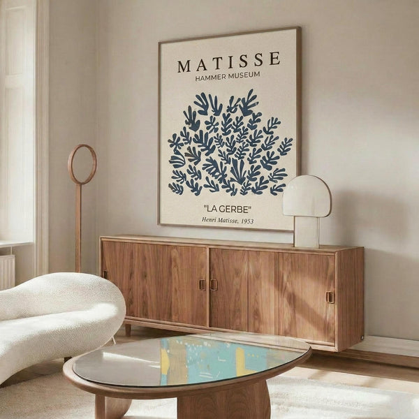 May include: Framed poster with blue botanical leaf cutout design on cream background, displayed above wooden sideboard in modern living room. White sculptural chair and glass-top coffee table with abstract art visible. Warm natural lighting creates shadows across neutral-toned space with minimalist decor.