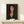 May include: Framed art print displaying a portrait of a woman with dark hair and elongated features in warm earth tones against a black background, styled in early 20th century modernist painting technique. The wooden-framed piece sits on a light wood shelf next to a small white wrapped gift with greenery, against a cream colored wall.
