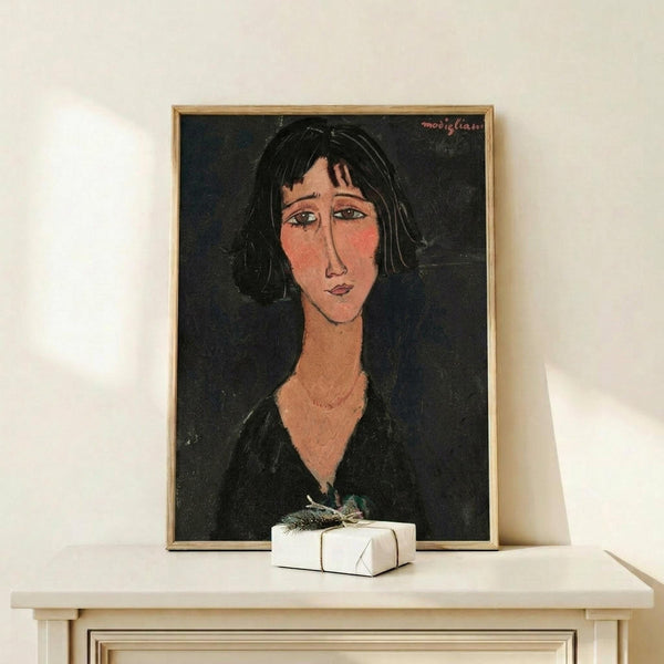 May include: Framed art print displaying a portrait of a woman with dark hair and elongated features in warm earth tones against a black background, styled in early 20th century modernist painting technique. The wooden-framed piece sits on a light wood shelf next to a small white wrapped gift with greenery, against a cream colored wall.