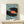 May include: Framed artwork showing a large black vinyl record with orange center label and turntable arm, surrounded by tiny people figures with shadows on light blue background. Natural wood frame displayed on light wood surface next to small wrapped gift box. Contemporary surreal art style with miniature scale contrast.