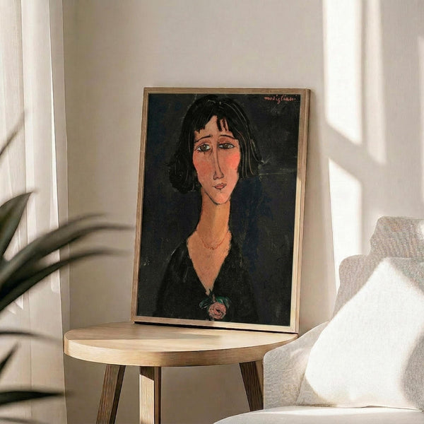 May include: Framed portrait print displayed on round wooden side table next to white upholstered chair with throw pillow. Artwork shows woman with dark hair, elongated face, and black dress against dark background in modernist style. Natural lighting creates soft shadows across white wall. Plant leaves visible in corner.