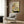 May include: Japanese woodblock print artwork hanging on wood-paneled wall above beige upholstered chair with geometric pillow. The vintage print features a striped cat drinking from an ornate ceramic bowl with palm tree and Japanese text, displaying warm earth tones of green, brown, and cream. Modern brass floor lamp and dark side table with open book complete the contemporary room setting.