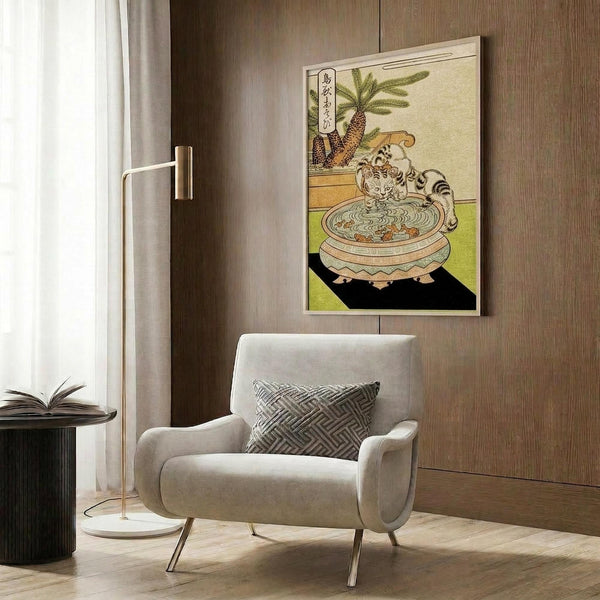 May include: Japanese woodblock print artwork hanging on wood-paneled wall above beige upholstered chair with geometric pillow. The vintage print features a striped cat drinking from an ornate ceramic bowl with palm tree and Japanese text, displaying warm earth tones of green, brown, and cream. Modern brass floor lamp and dark side table with open book complete the contemporary room setting.