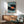 May include: Framed artwork showing a black vinyl record with orange center label surrounded by tiny people casting shadows on a light blue background. The print hangs above a wooden sideboard with white ceramic lamp in a mid-century modern living room with mint green upholstered chair and round coffee table. Natural sunlight creates dramatic shadows across white walls.