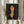 May include: Person in cream blouse holding framed art print featuring woman with dark bob haircut, elongated face, and black dress with small rose detail, painted in Modigliani's distinctive style with warm flesh tones against dark background. Dried wheat stalks visible on left side of neutral-toned room setting.