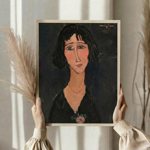 May include: Person in cream blouse holding framed art print featuring woman with dark bob haircut, elongated face, and black dress with small rose detail, painted in Modigliani's distinctive style with warm flesh tones against dark background. Dried wheat stalks visible on left side of neutral-toned room setting.