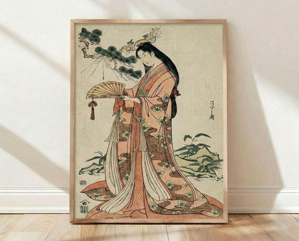 May include: Framed Japanese woodblock print artwork displayed on light wooden floor against white wall with natural lighting. Shows elegant woman in flowing orange and cream kimono with floral patterns holding decorative fan, surrounded by pine trees and foliage in traditional ukiyo-e style. Light wood frame complements the warm earth tones and delicate artistic details of the vintage-inspired reproduction print.