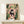 May include: Framed illustration of a black cat with green eyes sitting on a pink toilet while reading a newspaper, surrounded by lush green houseplants including trailing vines and potted plants. The artwork features a coral pink bathroom setting with botanical prints on the wall. Natural wood frame displayed on a light wooden surface beside a small white wrapped gift with greenery accent.