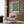 May include: Framed William Morris botanical print with intricate blue floral pattern on cream background, displayed on wood-paneled wall above beige upholstered armchair with geometric pillow. Scene includes brass floor lamp, dark round side table with open book, and natural lighting from window with curtains.