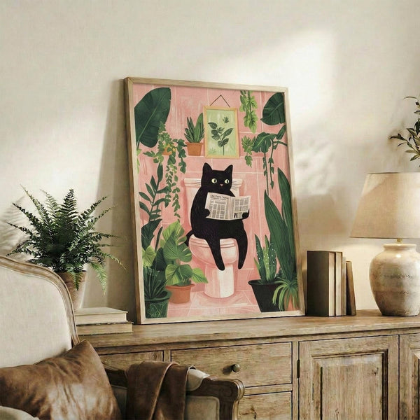 May include: Framed artwork showing a black cat with yellow eyes sitting on a pink toilet reading a newspaper, surrounded by lush green houseplants in terracotta pots against coral pink bathroom tiles. The illustration has a whimsical, flat design style with botanical prints on the wall. Displayed on a rustic wooden console table alongside fern plant, books, ceramic vase, and lamp in a cozy living room setting.