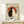 May include: Framed poster print showing two cats - one black, one white - sitting together against a large orange-red circle resembling a sun or moon, with Japanese text characters on either side. The artwork has a vintage, weathered appearance with cream and beige tones in an Asian-inspired style. The light wood frame sits on a pale wooden shelf with a small white wrapped gift box beside it, displayed against a white wall with soft natural lighting.