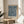 May include: Blue and cream William Morris floral pattern poster in light wood frame, displayed on round wooden side table next to beige upholstered chair. Sunlight streams across white walls creating geometric shadows. Green plant leaves visible in corner. The intricate botanical design features symmetrical flowers, leaves, and vines in navy blue on cream background.
