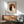 May include: Framed artwork showing two cats in yin yang pose against orange circle background with Japanese characters, displayed above wooden sideboard with table lamp in mid-century modern living room setting with sage green chair and natural lighting creating shadows on white wall