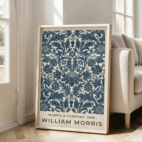 May include: A framed poster featuring an intricate blue and cream botanical pattern with symmetrical floral motifs, grape clusters, and ornate leaves. The artwork displays "MORRIS & COMPANY, 1866" and "WILLIAM MORRIS" text at the bottom. The light wooden frame leans against a white wall next to a beige upholstered sofa in a bright, sunlit room with white trim and hardwood floors.