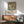 May include: Framed botanical exhibition poster featuring large cream peony flower with green foliage on vintage-style background, displayed above wooden sideboard in mid-century modern living room with mint green upholstered chair, ceramic table lamp, and natural lighting casting shadows on white wall.