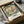 May include: Vintage botanical poster featuring a large cream and coral peony flower with green foliage, displayed in black frame within cardboard packaging box surrounded by brown paper wrapping materials on white tissue paper surface.