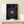 May include: A framed navy blue poster with gold text displaying song lyrics arranged in a spiral pattern around a central vinyl record image, positioned on a light wood shelf next to a small white wrapped gift with twine and greenery, against a cream colored wall.