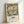 May include: Framed vintage-style exhibition poster featuring large cream and coral botanical flower with detailed petals and stamens, surrounded by teal and green foliage. Text below reads "Cotton Print Exhibition" with Underground station logo and South Kensington location details. Light wood frame against white wall, soft natural lighting creating subtle shadows.