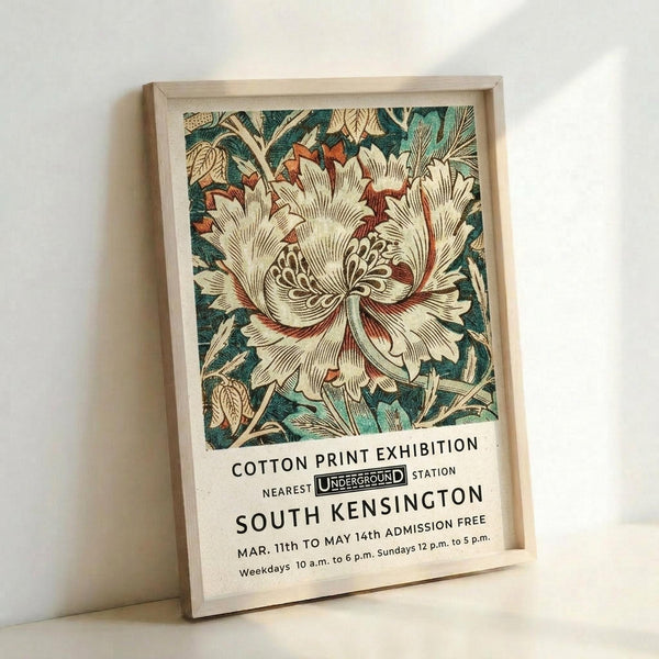 May include: Framed vintage-style exhibition poster featuring large cream and coral botanical flower with detailed petals and stamens, surrounded by teal and green foliage. Text below reads "Cotton Print Exhibition" with Underground station logo and South Kensington location details. Light wood frame against white wall, soft natural lighting creating subtle shadows.