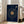 May include: Framed navy blue poster with golden vinyl record design featuring song lyrics arranged in curved text around a central orange and black record. Brass-colored tonearm extends from top right. White text at top shows placeholder song and artist name, with names Oliver & Meaghan at bottom. Light wood frame leans against white wall beside beige upholstered chair in bright, minimalist living room with white walls and large windows.