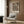 May include: Vintage botanical poster with dark navy background featuring ornate floral pattern in coral, cream and gold tones, displayed in light wood frame on textured wood panel wall above modern cream armchair with geometric pillow, brass floor lamp and round side table with open book nearby