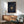 May include: Navy blue poster with spiral text design and vinyl record center, displayed in wooden frame above mid-century modern credenza. Scene shows sage green upholstered chair with wooden arms, small white table lamp, and round coffee table in sunlit room with white walls.