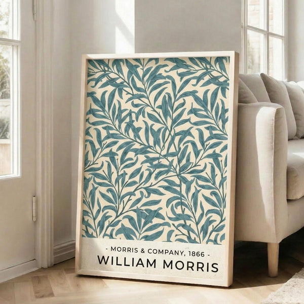 May include: Framed botanical art print featuring William Morris's signature willow leaf pattern in teal green on cream background, displayed on wooden floor against white walls next to beige upholstered armchair. The vintage-style poster shows flowing, intricate leaf motifs with Morris & Company, 1866 text at bottom. Natural lighting streams through nearby windows onto the hardwood floors.