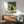 May include: Framed poster displaying a black cat with glowing yellow eyes sitting beside a vase of orange and white flowers, with pizza slices in a box on a table. The artwork features green and cream tones in a vintage illustration style. Shown hanging above a mid-century modern wooden credenza with a white ceramic lamp, next to a teal upholstered armchair with wooden frame. Sunlight streams across white walls creating geometric shadows.