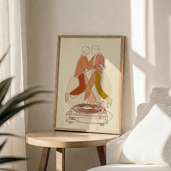 May include: Framed poster print showing two stylized figures in warm orange, coral and yellow tones dancing above a vintage turntable, displayed on a round wooden side table next to a beige upholstered chair with white throw pillow, soft natural lighting creating shadows on neutral wall background with plant leaves visible