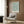 May include: Framed botanical print with blue-green willow leaf pattern on cream background, displayed above a beige upholstered armchair with geometric pillow. Modern living room setting features wood paneling, brass floor lamp, and dark side table with open book.