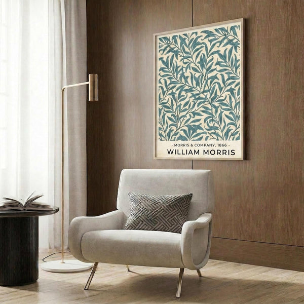 May include: Framed botanical print with blue-green willow leaf pattern on cream background, displayed above a beige upholstered armchair with geometric pillow. Modern living room setting features wood paneling, brass floor lamp, and dark side table with open book.