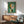 May include: Abstract geometric artwork with green background featuring circular and angular shapes in orange, white, pink, and black, displayed in wooden frame above mid-century modern credenza with white ceramic lamp, alongside teal upholstered armchair in sunlit room with white walls