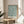 May include: Framed botanical print with teal blue willow branch pattern on cream background, displayed on round wooden side table next to beige upholstered chair. Natural lighting creates soft shadows across neutral-toned room with plant leaves visible in corner. Print features flowing organic leaf design in vintage Arts and Crafts style with text attribution at bottom.