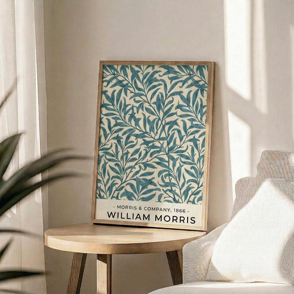 May include: Framed botanical print with teal blue willow branch pattern on cream background, displayed on round wooden side table next to beige upholstered chair. Natural lighting creates soft shadows across neutral-toned room with plant leaves visible in corner. Print features flowing organic leaf design in vintage Arts and Crafts style with text attribution at bottom.