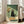 May include: Framed artwork showing a black cat with large white eyes sitting beside a glass vase of orange and cream flowers, with an open pizza box containing four slices on a wooden surface. The illustration has a vintage, textured appearance with green tones. The wooden-framed print leans against a white wall next to a beige upholstered sofa in a bright living room with large windows.
