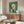 May include: Abstract modernist artwork with geometric shapes in green, orange, and pale colors displayed above a wooden sideboard in a minimalist living room. The framed print features circular and angular forms against a textured green background. White upholstered chair and glass coffee table complete the mid-century modern setting with natural lighting.