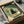 May include: Framed art print in black frame being unboxed from cardboard packaging with protective wrapping. The artwork shows a black cat with wide yellow eyes sitting next to a vase of orange and white flowers, with pizza slices on an open box below. The illustration has a vintage poster style with muted green background and warm tones. Frame appears medium-sized relative to the shipping box.