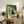 May include: Framed artwork displaying a black cat with white eyes sitting beside a vase of orange and white flowers, with a pizza box containing four slices below. The illustration features green geometric wall sections in the background. The wooden-framed piece is styled on a rustic wooden console table alongside books, a ceramic vase, and a fern plant in a cozy living room setting with beige furniture.